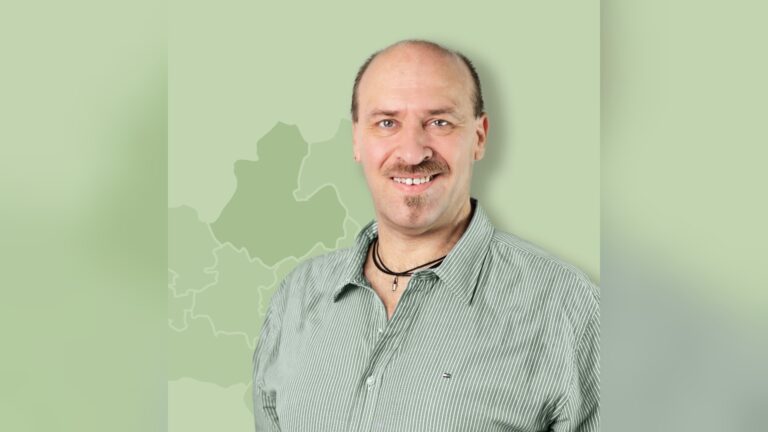 Michael Böttcher, ein lächelnder Mann mit Schnurrbart und Glatze in gestreiftem Hemd vor einer grünen Karte