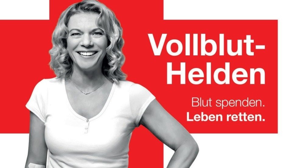 Lächelnde Frau mit Pflaster am Arm vor rotem Hintergrund mit DRK-Kreuz und dem Text Vollblut-Helden, Blut spenden