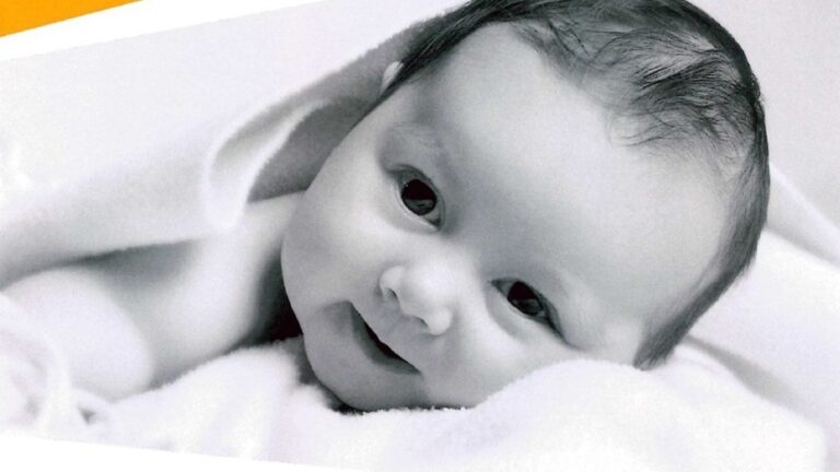 Kleines Baby mit dunklem Haar liegt auf einem weißen Tuch, lächelt leicht und schaut in die Kamera