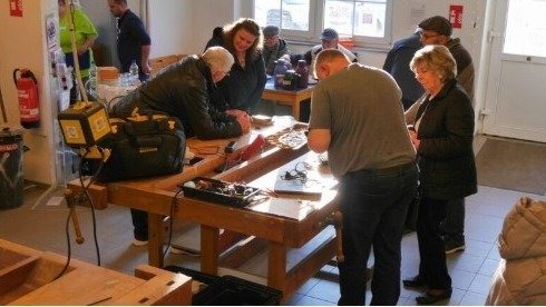 Eine Gruppe Menschen repariert gemeinsam verschiedene Gegenstände an einer langen Holzwerkbank in einer Werkstatt