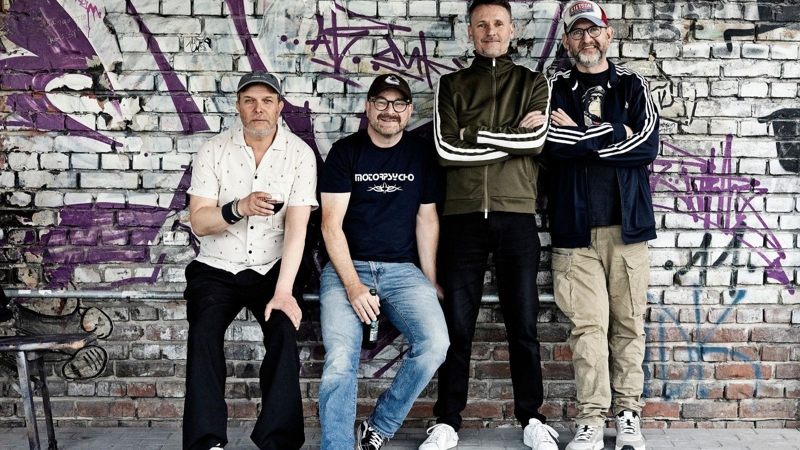 Vier Bandmitglieder der Seltaebs posieren vor einer graffitibesprühten Ziegelwand, zwei sitzen, zwei stehen mit