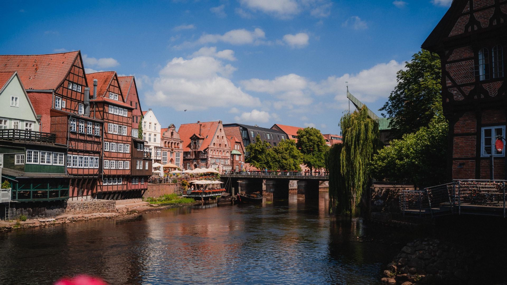 Fachwerkhäuser und Restaurants am Lüneburger Stintmarkt am Flussufer mit einer Brücke und Menschen unter blauem Himmel