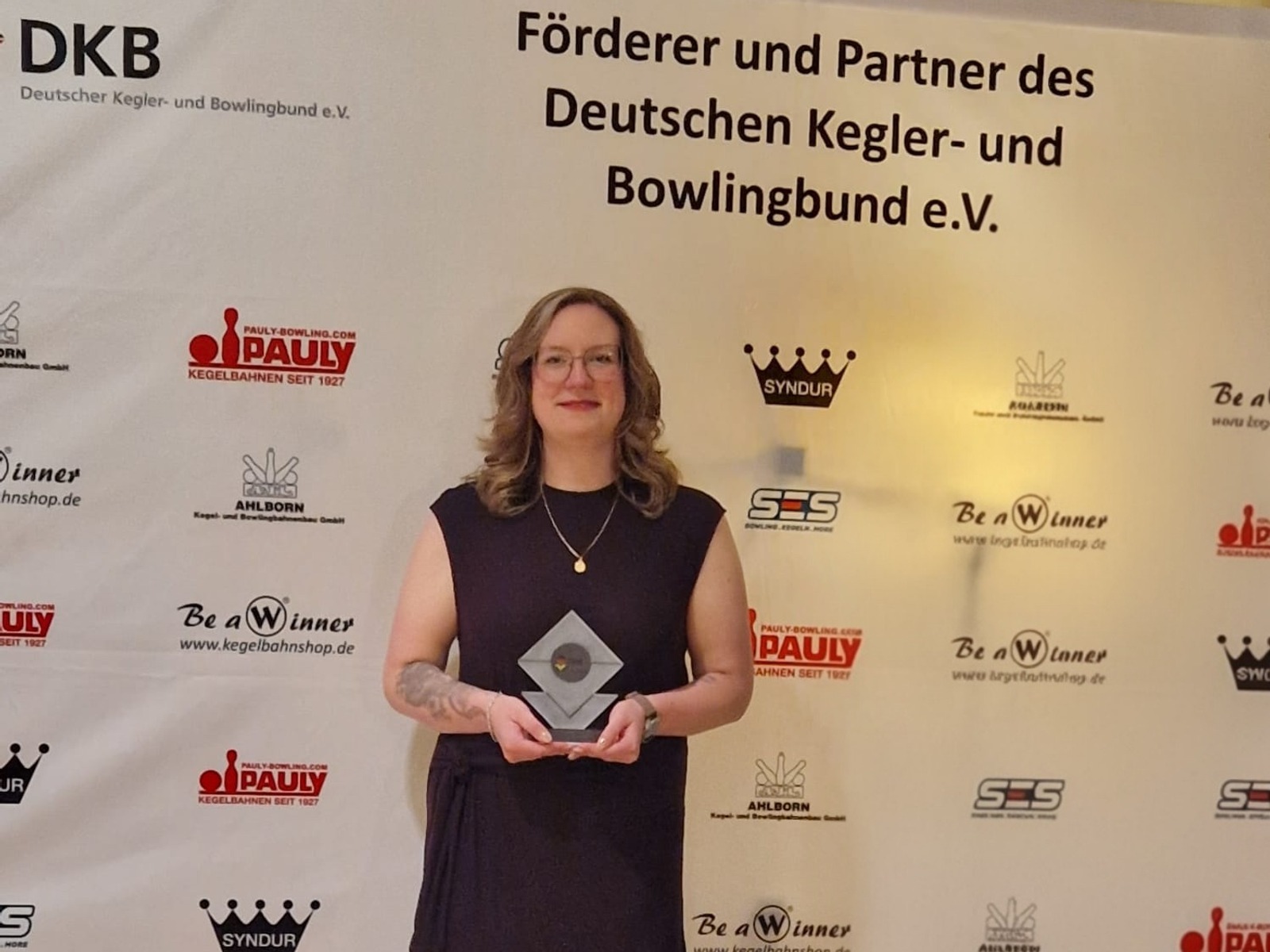 Christina Rähse mit Brille und dunklem Kleid steht vor einer Sponsorenwand und hält eine gläserne Auszeichnung