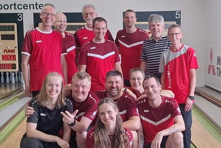 Eine Gruppe lachender Sportkegler in roten Trikots posiert auf einer Kegelbahn mit Pins und Anzeigetafel
