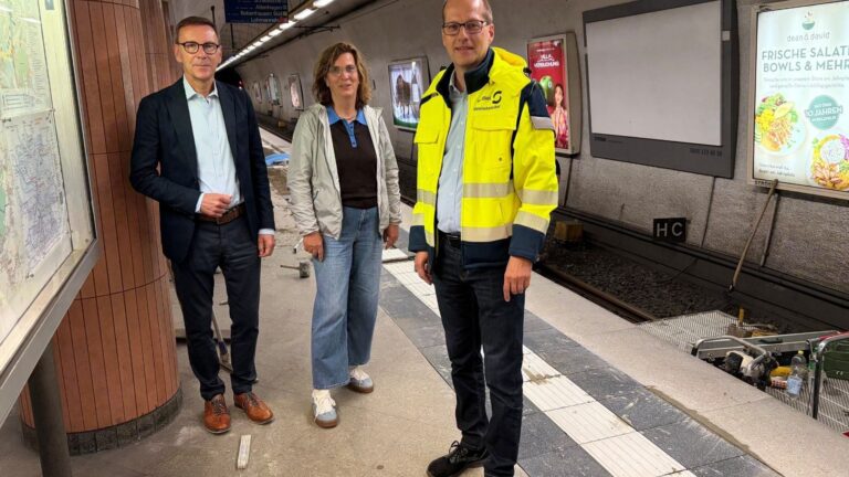 Drei Personen, ein Mann im Anzug, eine Frau in Jeans und ein Mann in gelber Warnweste, auf einem Bahnsteig im