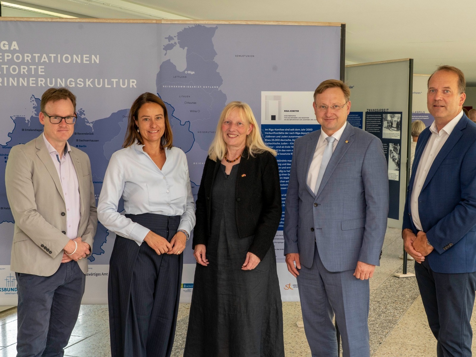 Vier Männer und eine Frau stehen lächelnd vor einer Ausstellung zu Deportationen und dem Riga-Komitee