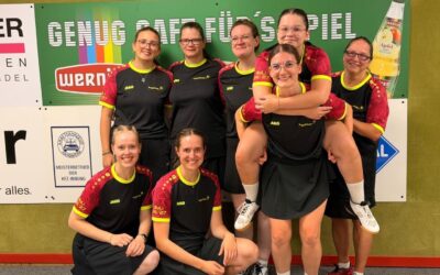 Acht lächelnde Frauen eines Kegelteams in schwarzen Trikots, eine trägt eine andere huckepack, vor einer grünen Wand