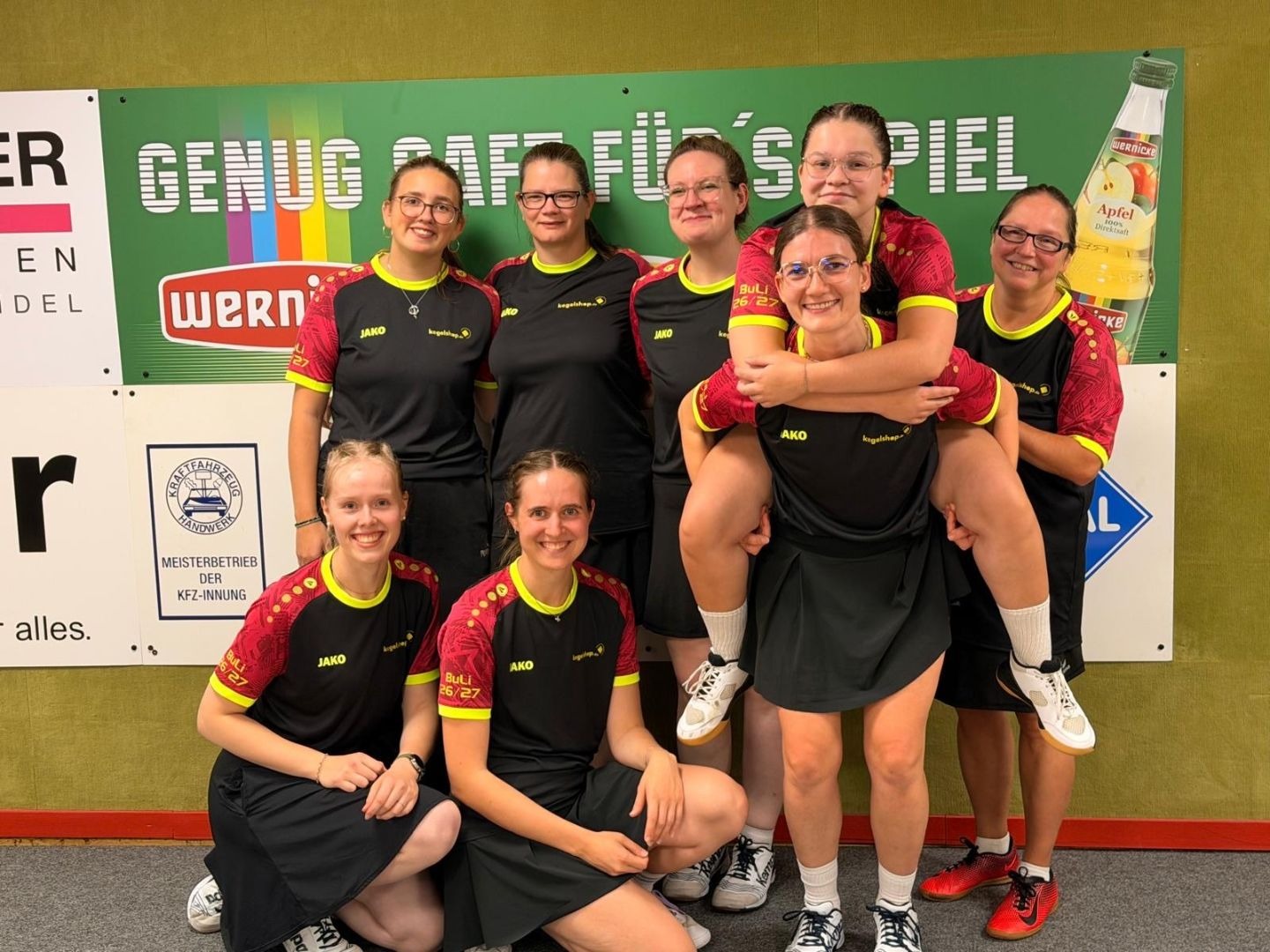Acht lächelnde Frauen eines Kegelteams in schwarzen Trikots, eine trägt eine andere huckepack, vor einer grünen Wand