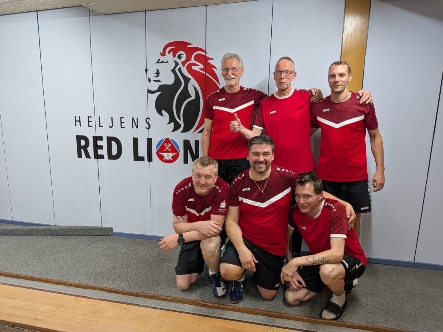 Sechs Männer in roten Trikots und schwarzen Hosen, drei stehend, drei kniend, vor dem roten Löwen-Logo