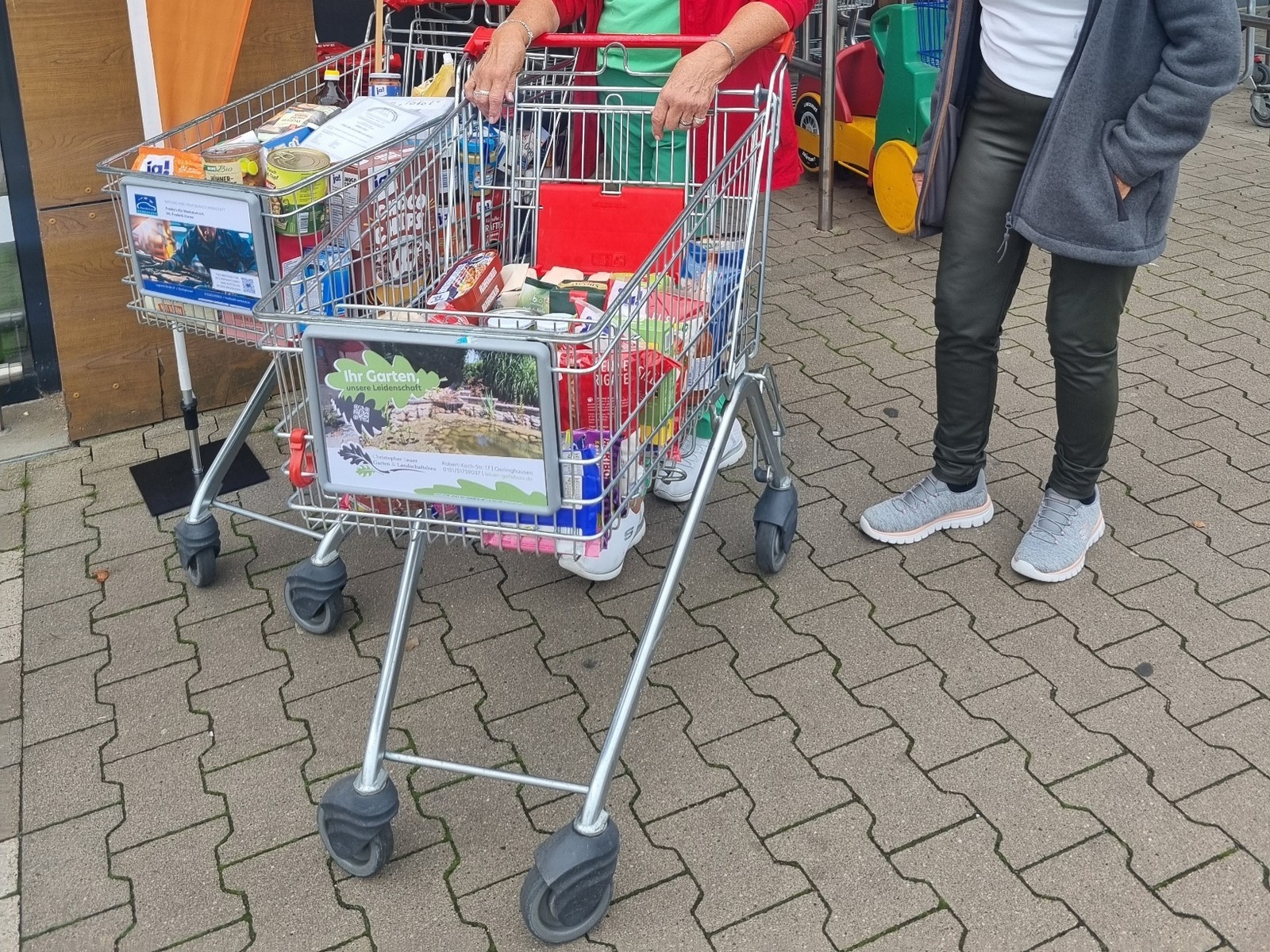 Zwei lächelnde Frauen mit einem vollgepackten Einkaufswagen für die Tafeln vor einem Rewe-Supermarkt