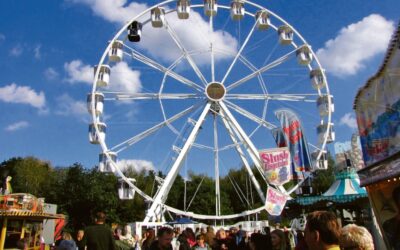 Belebter Kirmesplatz mit einem großen weißen Riesenrad, Buden und vielen Besuchern unter blauem Himmel