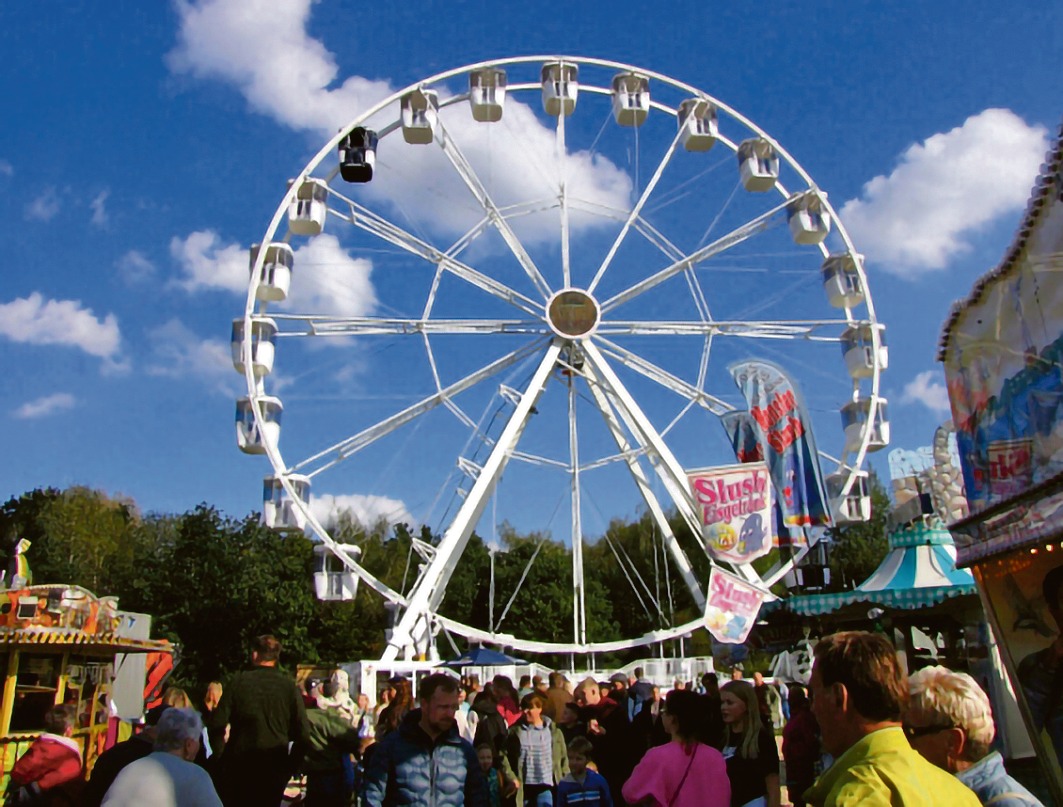 Belebter Kirmesplatz mit einem großen weißen Riesenrad, Buden und vielen Besuchern unter blauem Himmel