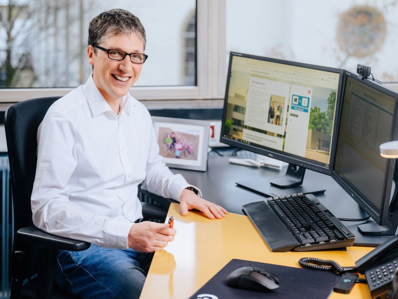 Lächelnder Mann mit Brille und weißem Hemd sitzt an einem Schreibtisch mit zwei Monitoren, Telefon und Familienfoto