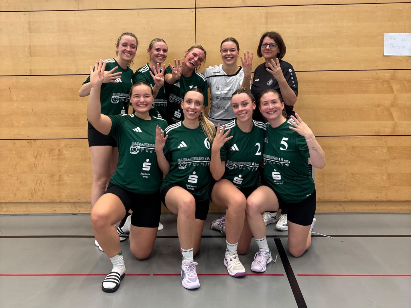 Lachendes Frauen-Sportteam aus Helpup mit Trainerin in Sporthalle, viele zeigen vier Finger