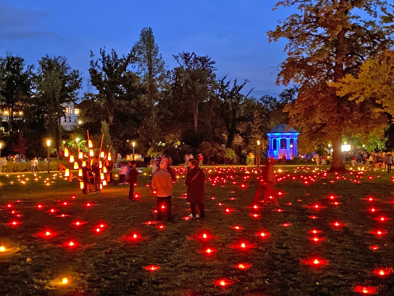 Dunkler Park mit Menschen, die rote Bodenlichter, eine hohe Laternenkonstruktion und einen blau leuchtenden Pavillon