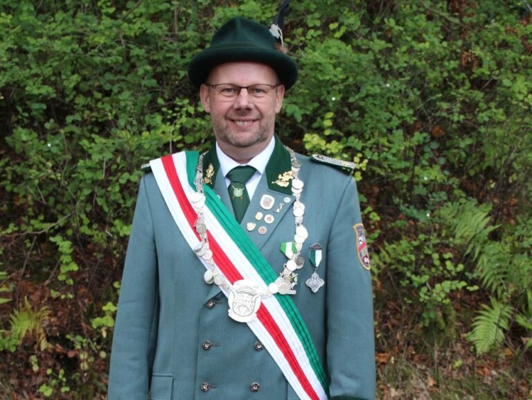 Mann in grüner Schützenuniform mit Orden und Schützenkette lächelt vor Grünpflanzen