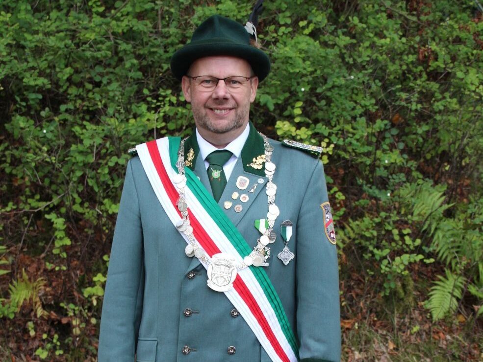 Mann in grüner Schützenuniform mit Orden und Schützenkette lächelt vor Grünpflanzen