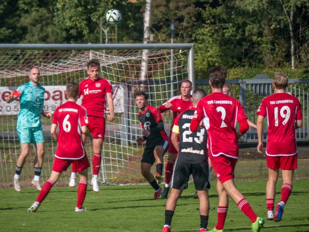 Roter Fußballspieler kickt Ball auf grünem Rasen, ein schwarzer Spieler läuft daneben, weitere Spieler und Werbebanden