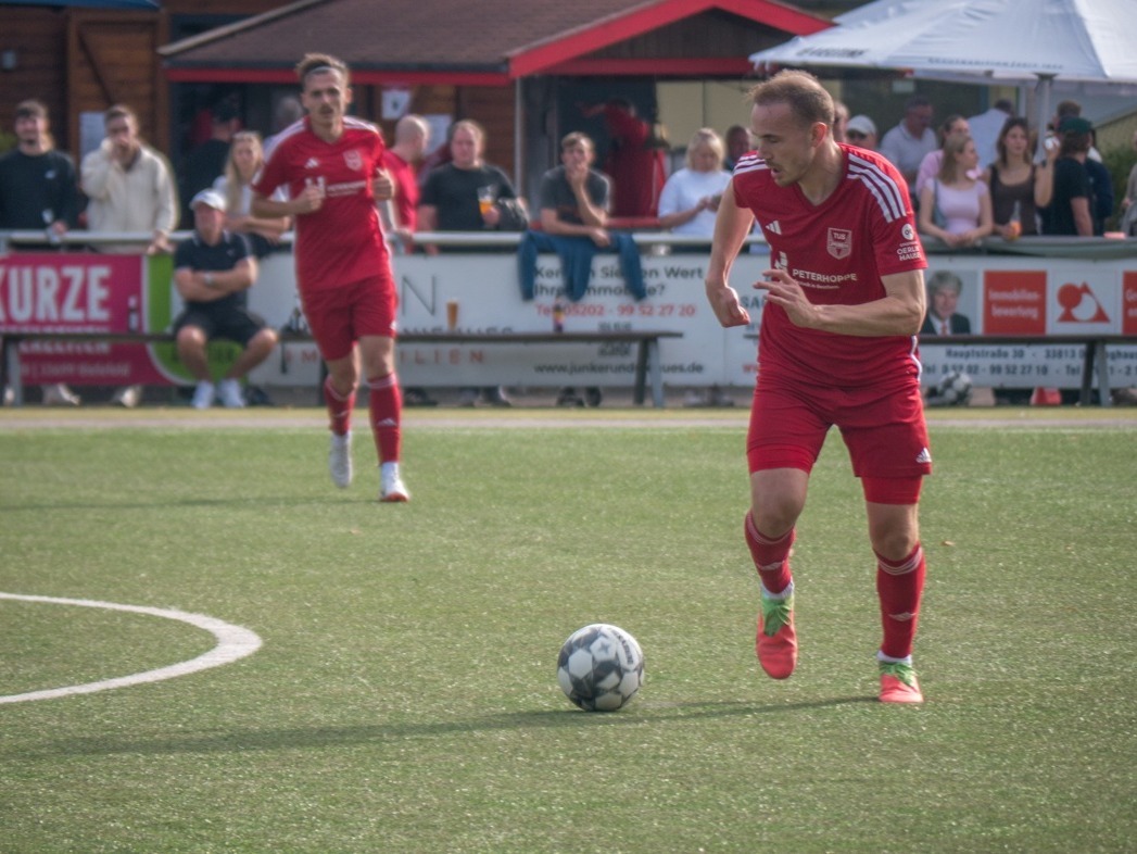 Ein Fußballspieler in rotem Trikot läuft auf einem grünen Rasenfeld neben einem Fußball, ein weiterer Spieler folgt im
