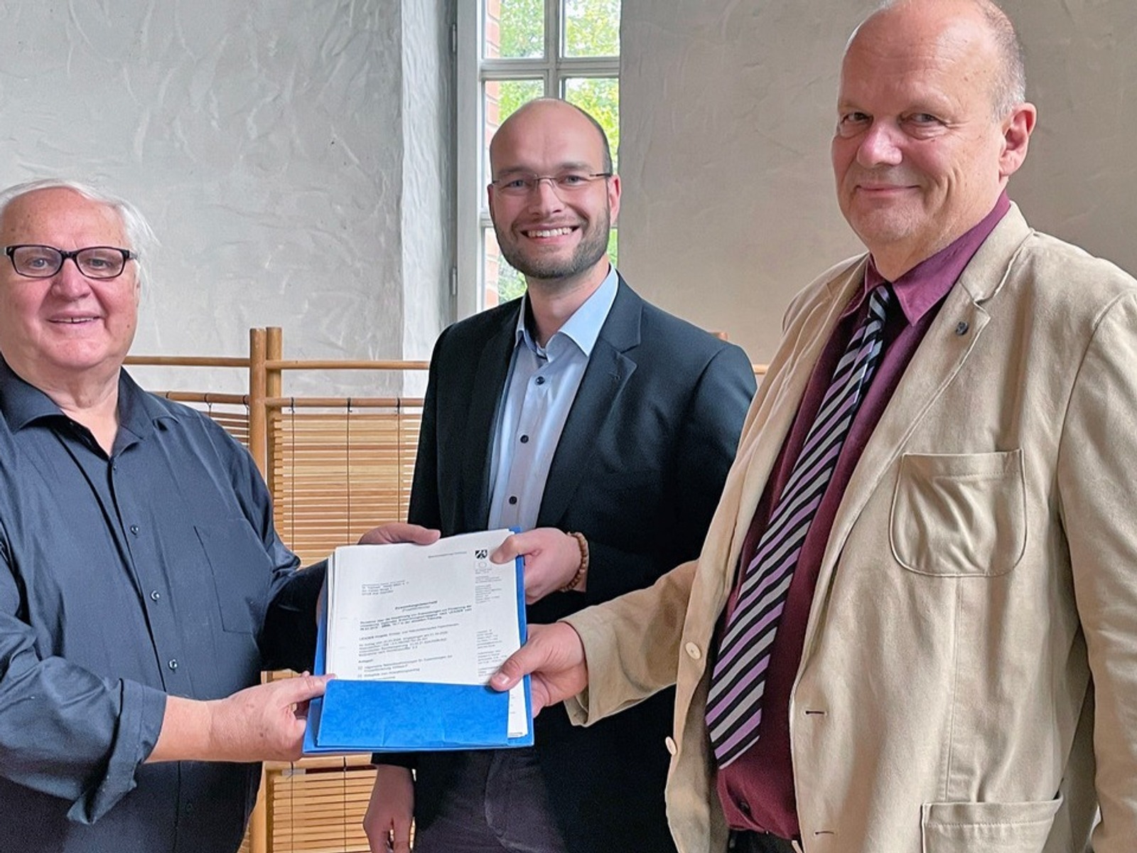 Drei lächelnde Männer überreichen einen Förderbescheid in einem Besprechungsraum