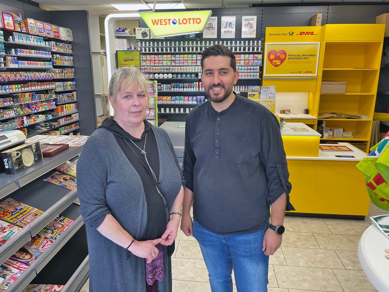 Eine blonde Frau und ein bärtiger Mann stehen lächelnd in einem Lotto- und Postladen vor Regalen