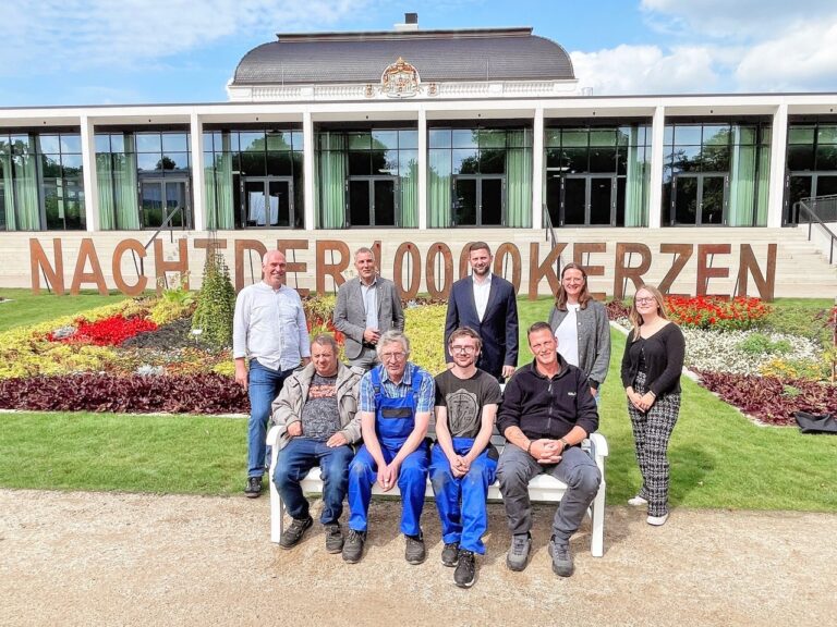 9 Personen stehen und sitzen vor einem Gebäude und einem Blumenbeet mit dem Schriftzug "Nacht der 1.000 Kerzen