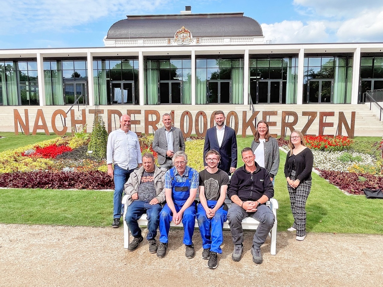 9 Personen stehen und sitzen vor einem Gebäude und einem Blumenbeet mit dem Schriftzug "Nacht der 1.000 Kerzen