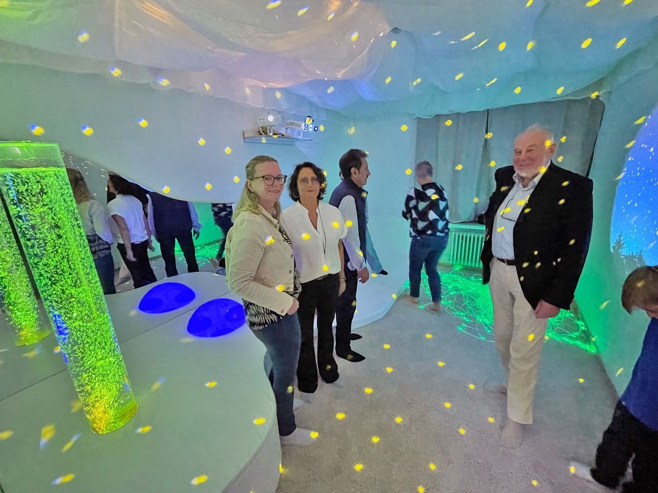 Personen in einem Snoezelraum mit grünen Blasensäulen, blauen Leuchtdiscs, gelben Lichtpunkten und Lichtprojektionen