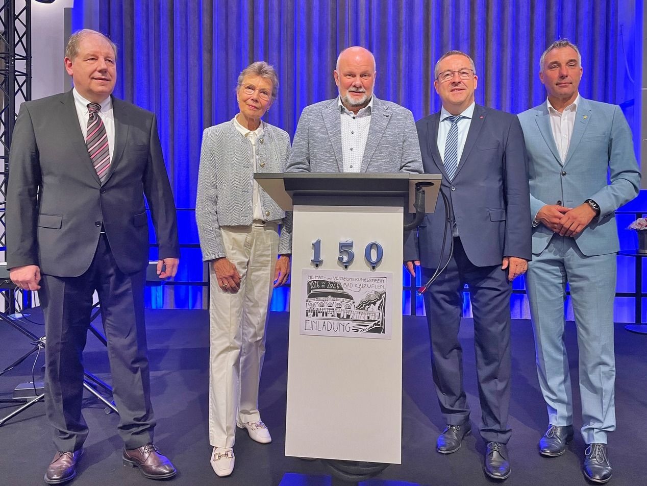 Vier Männer und eine Frau stehen auf einer Bühne vor einem Rednerpult mit einem 150-Jubiläums-Logo
