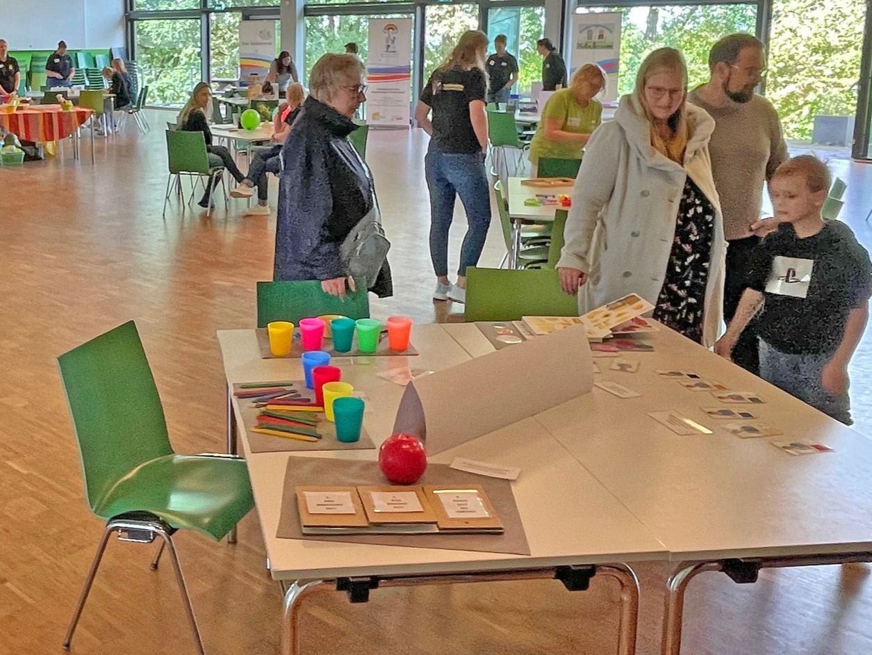 Personen, darunter eine Familie mit Kind, an Tischen mit bunten Bechern, Stiften und Karten in einer hellen Halle