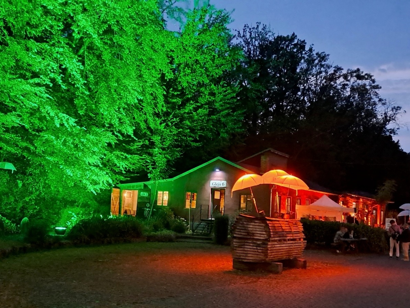 Abendliche Szene mit grün beleuchteten Bäumen, dem Gebäude Gleis 6, orangen Lichtinstallationen und vielen Personen