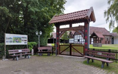 Eingangsbereich des Heimathofs Leopoldshöhe mit Holztor, Infotafel, zwei Bänken und traditionellen Gebäuden im