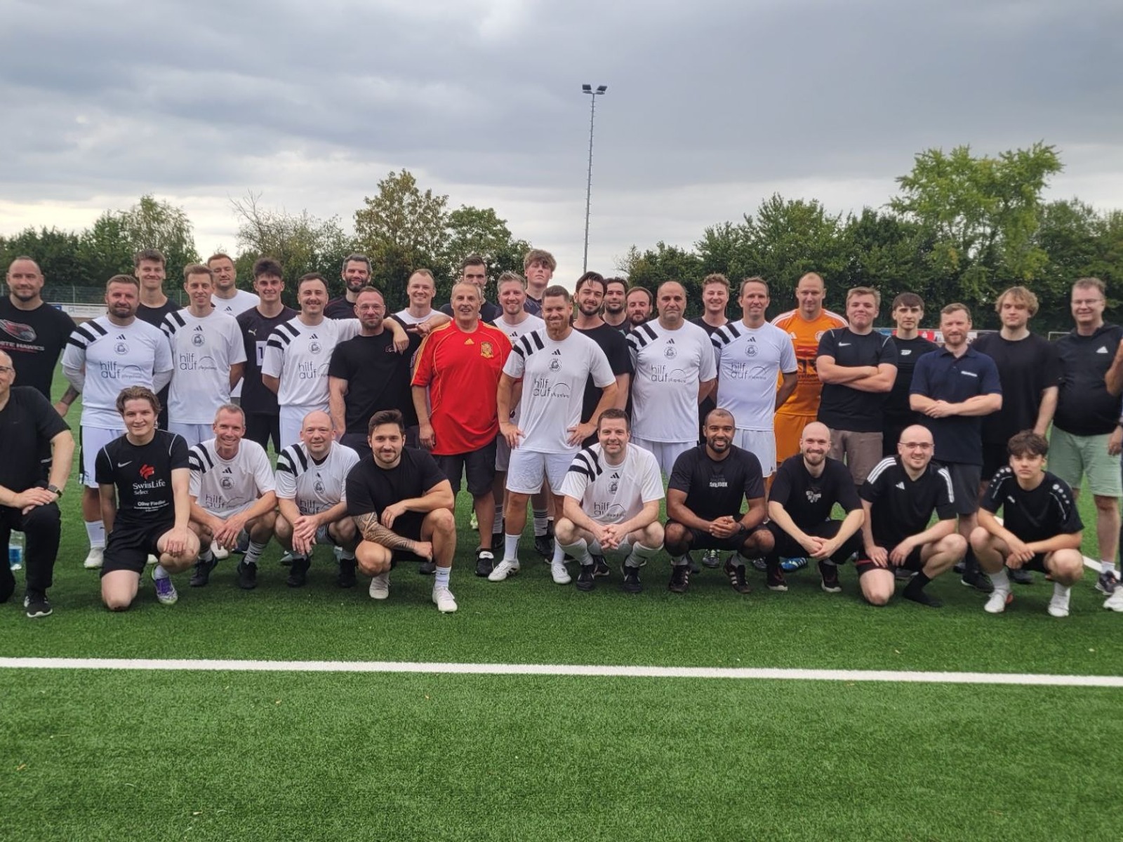 Große Gruppe Männer in Sportkleidung steht lächelnd auf grünem Kunstrasenplatz an einer weißen Linie