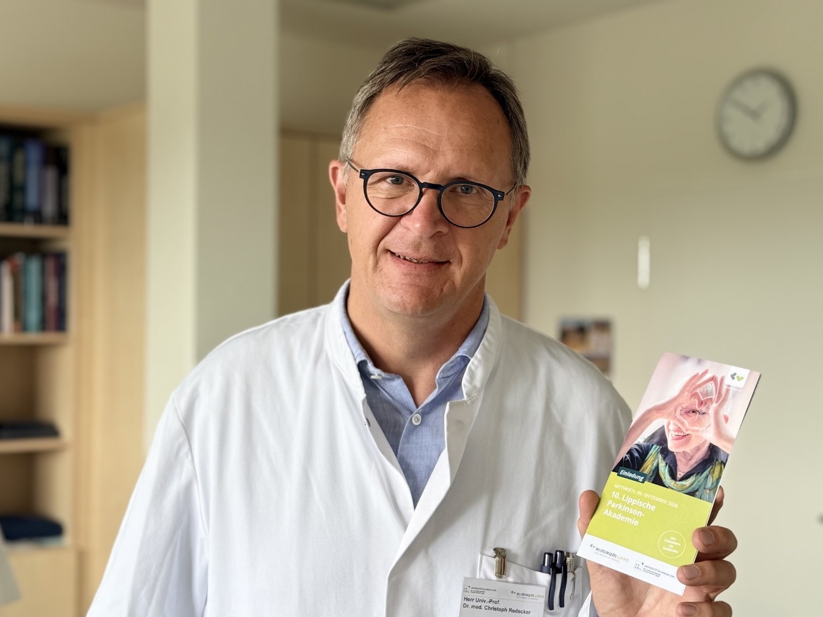 Mann in weißem Kittel und Brille hält Broschüre zur 10. Lippischen Parkinson-Akademie