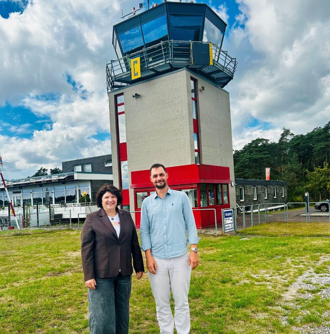 Landtagsabgeordnete Ellen Stock und ein Mann stehen vor einem Fluglotsenturm und einem Gebäude der