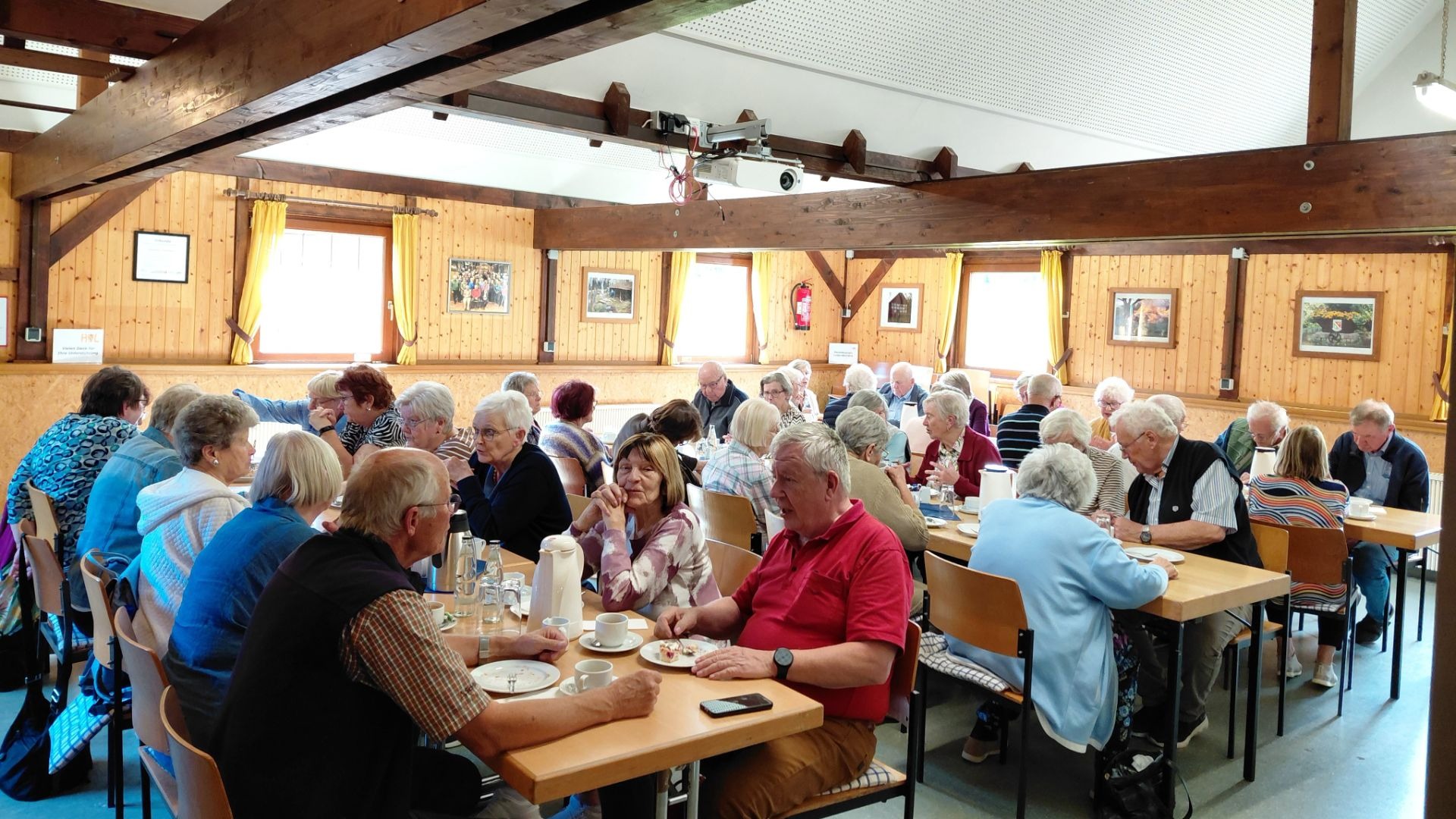 Zahlreiche Senioren sitzen in einem holzgetäfelten Raum an Tischen, trinken Kaffee und unterhalten sich lebhaft