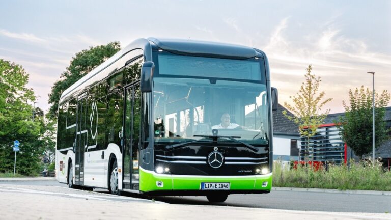Schwarz-weiß-grüner Stadtbus mit einem Fahrer am Steuer auf einer Straße vor Bäumen und Gebäuden