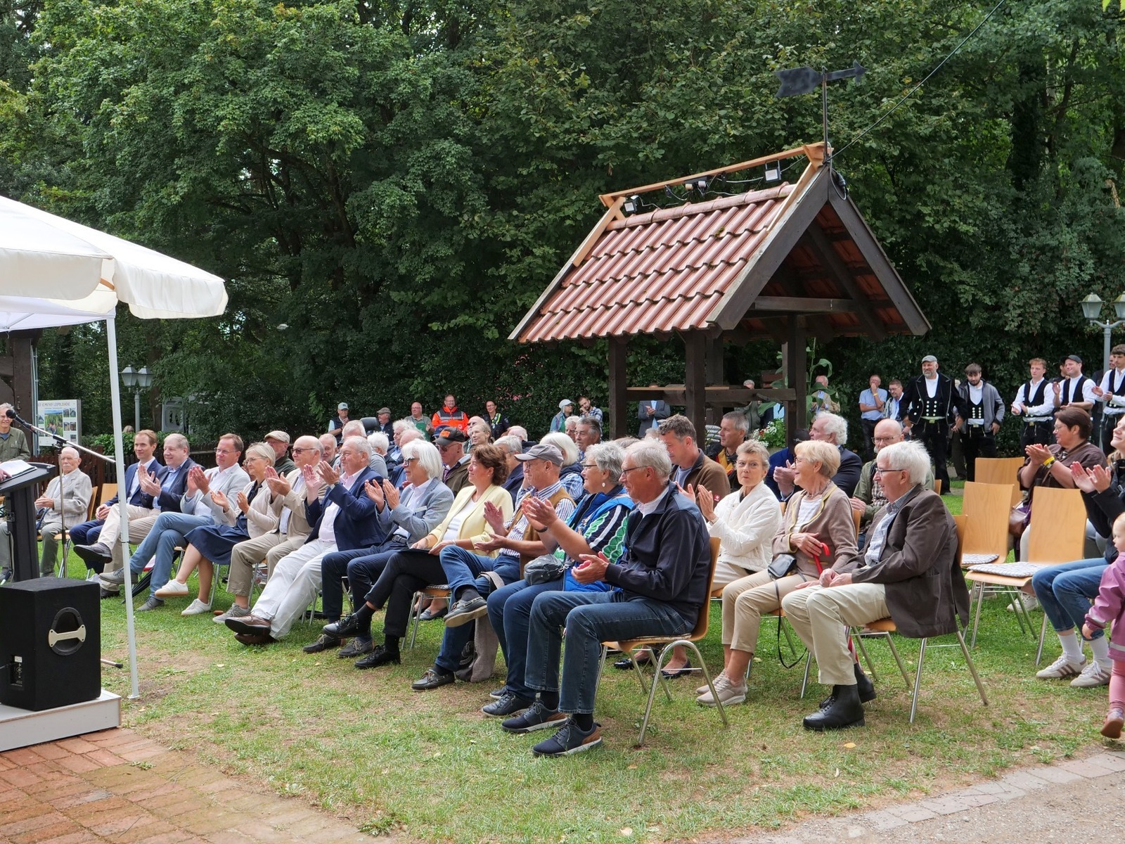 Redner spricht auf einer Bühne vor applaudierendem Publikum, Musikern und Trachtengruppe bei einem Fest im Grünen