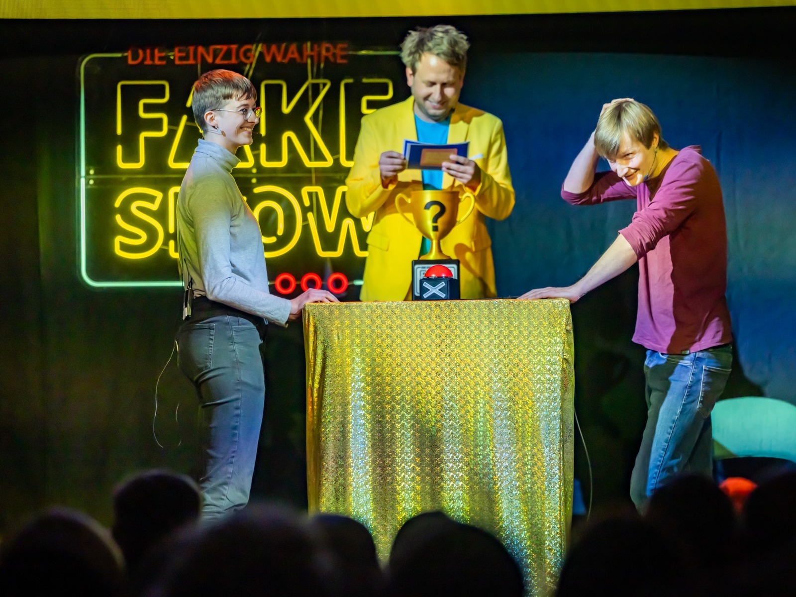 Drei Personen auf der Bühne einer Quizshow: Ein Moderator und zwei Kandidaten vor einem Leuchtschriftzug "Fake Show