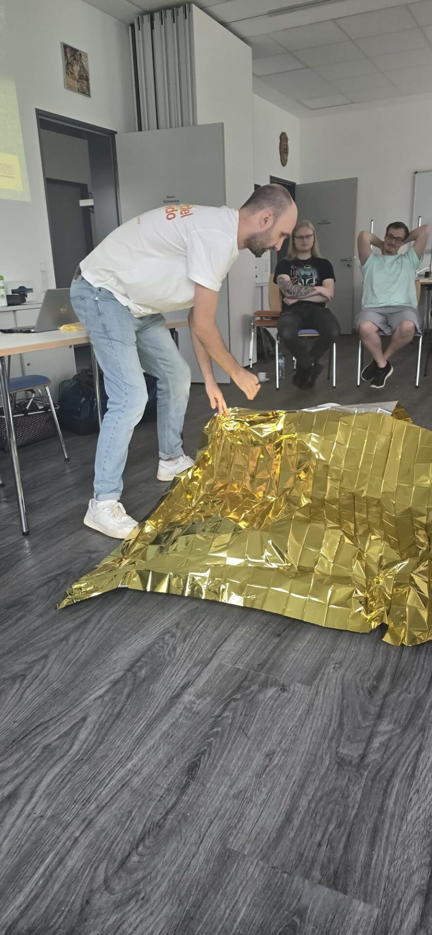 Ein Mann zeigt im Erste-Hilfe-Kurs die Anwendung einer goldenen Rettungsdecke vor drei sitzenden Teilnehmern