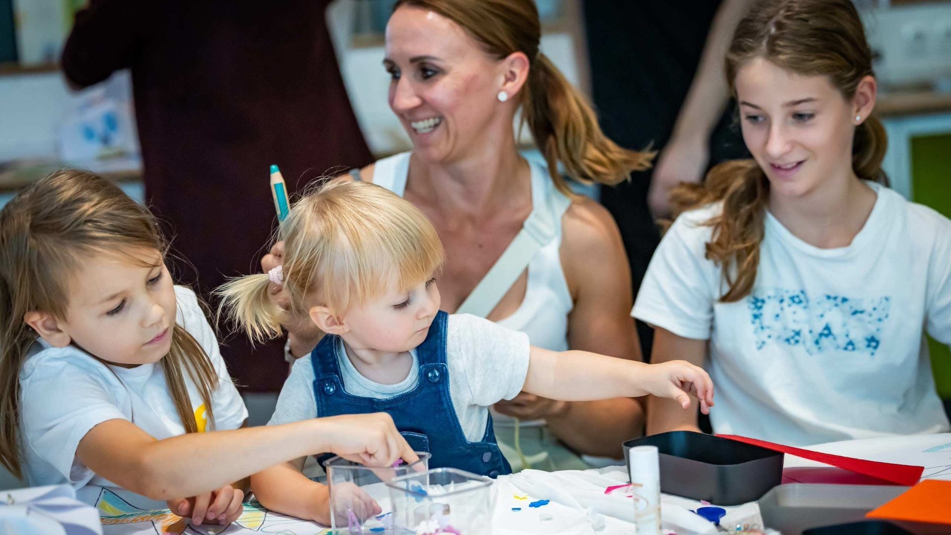 Zwei kleine Mädchen und eine Jugendliche basteln zusammen mit einer lachenden Frau an einem Tisch