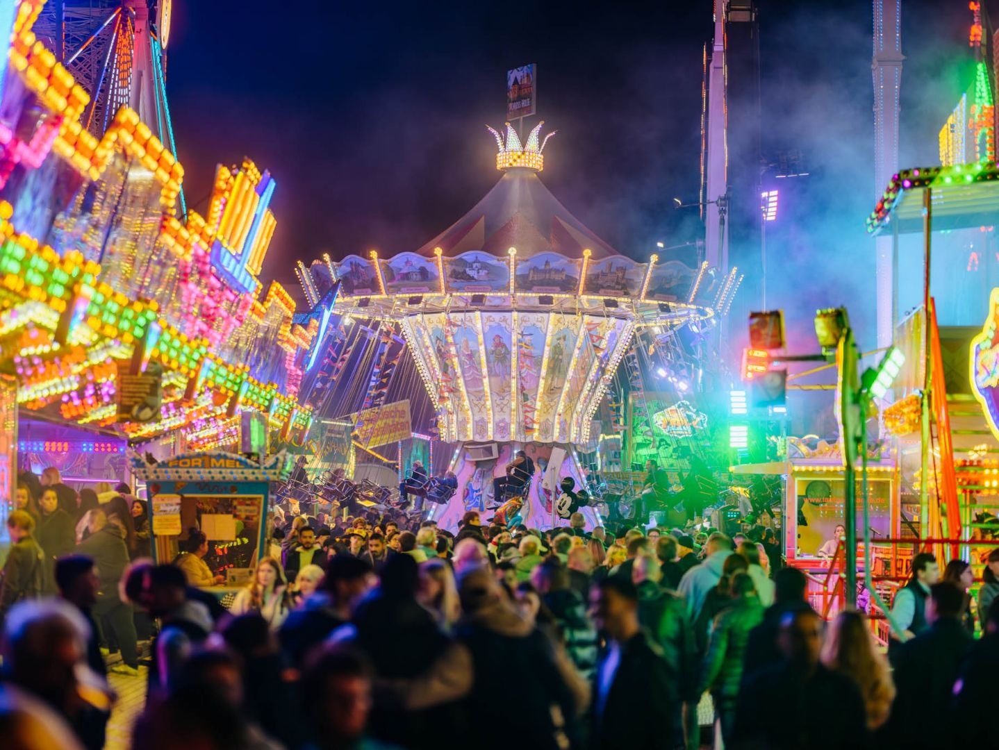 Menschenmenge auf einem nächtlichen Volksfest mit vielen bunten, beleuchteten Fahrgeschäften und einem Kettenkarussell