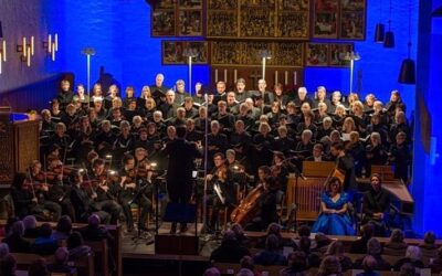 Großer singender Chor und spielendes Orchester in blau beleuchteter Kirche vor Publikum, mit Solisten und Dirigent