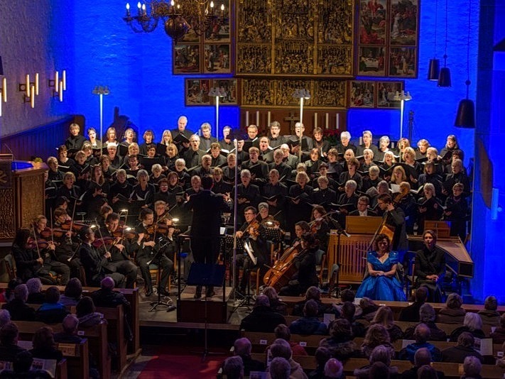 Großer singender Chor und spielendes Orchester in blau beleuchteter Kirche vor Publikum, mit Solisten und Dirigent