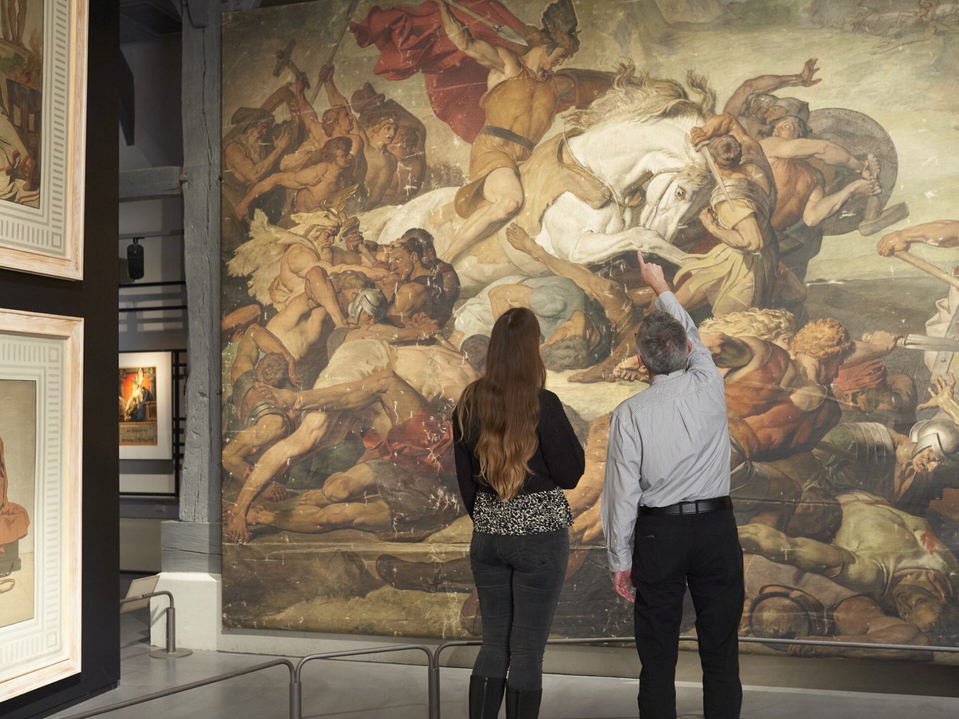 Zwei Personen von hinten betrachten ein großes Schlachtengemälde im Museum, ein Mann zeigt auf Details