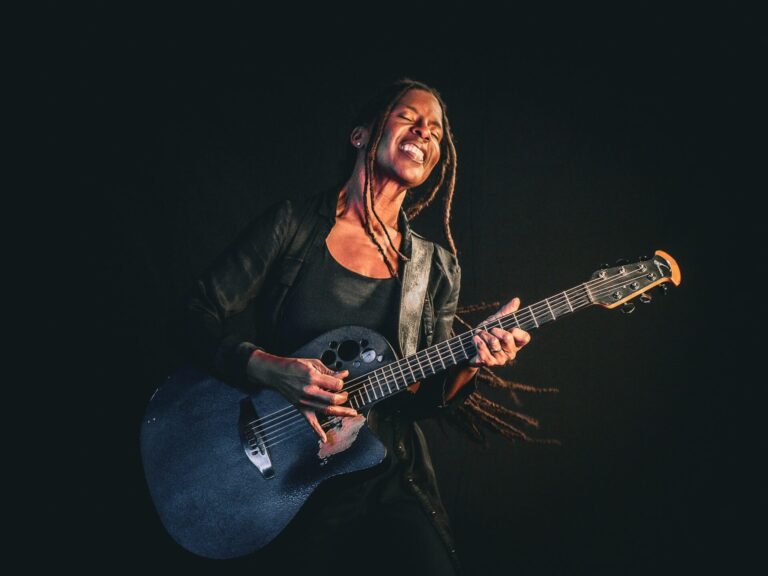 Judy Bailey spielt mit geschlossenen Augen und geöffnetem Mund eine dunkelblaue Akustikgitarre auf dunklem Hintergrund