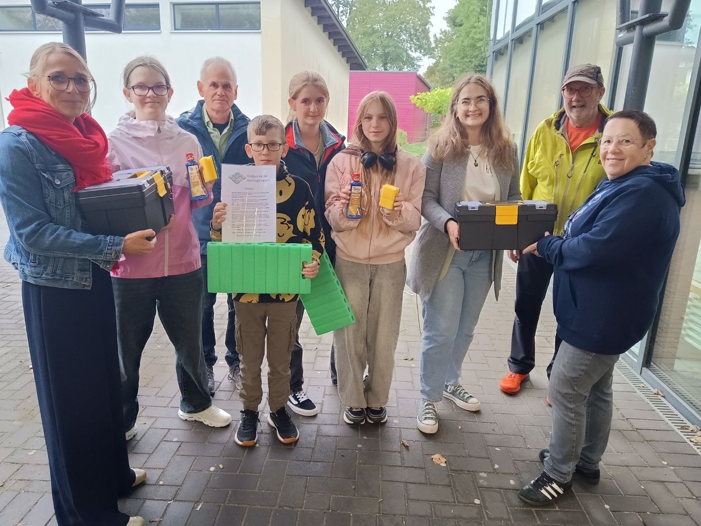 Acht lächelnde Personen, darunter Schüler, stehen draußen mit Putzmitteln, Werkzeugkästen, Knieschonern und einem