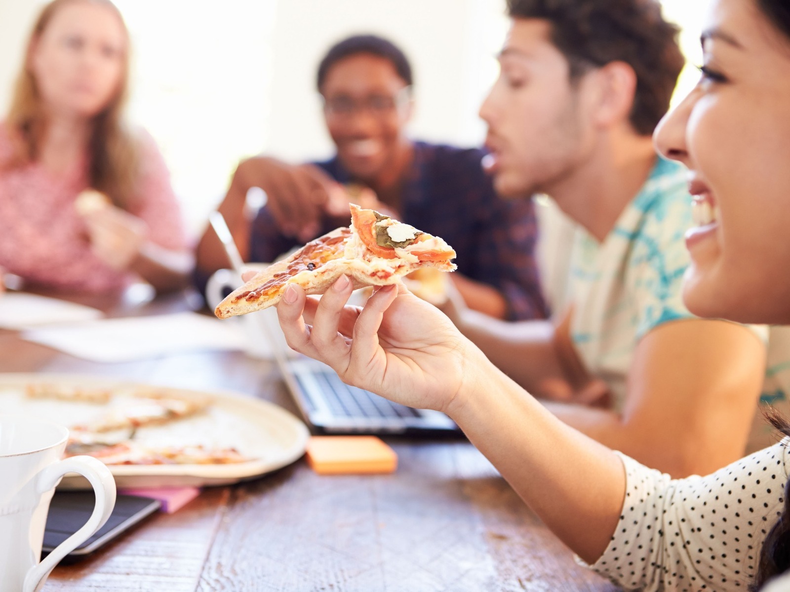 Eine lachende Frau hält ein Pizzastück vor dem Mund, im Hintergrund essen weitere Menschen an einem Tisch