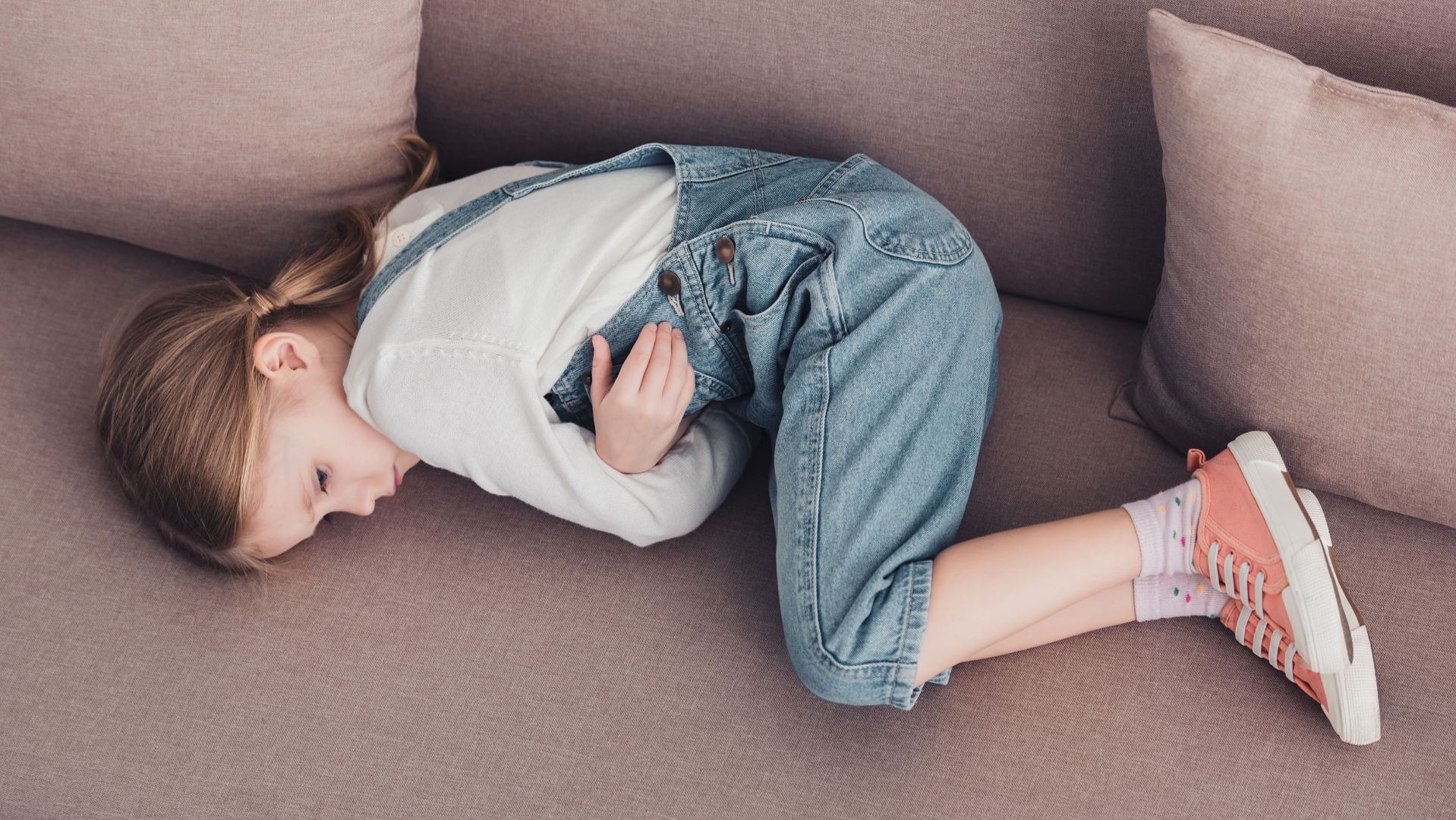 Kleines Mädchen mit Zopf in blauer Latzhose liegt gekrümmt auf der Seite auf einem beigen Sofa