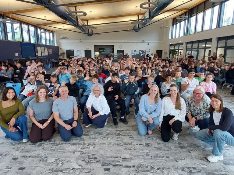 Viele winkende Fünftklässler und lächelnde Erwachsene sitzen in einer großen Schulaula für ein Gruppenfoto